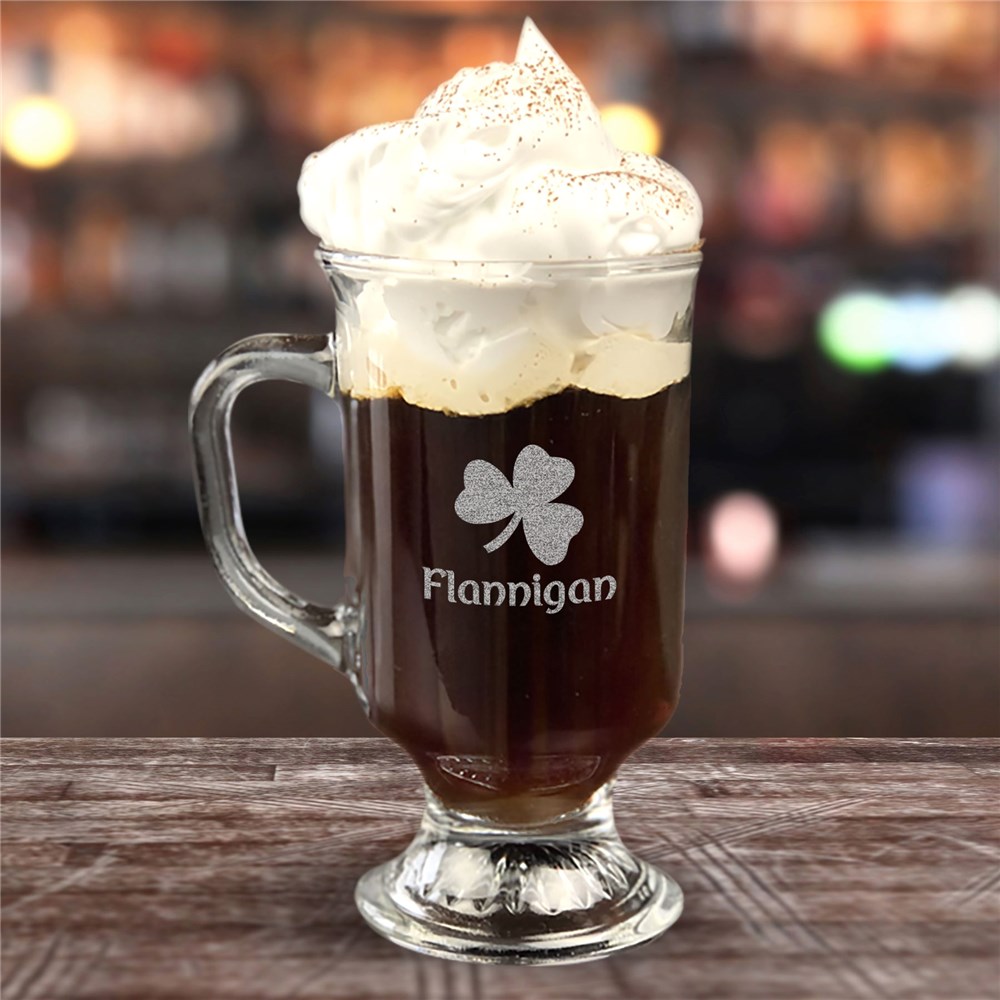 A clear glass mug with a handle, filled with coffee topped with whipped cream, features an engraved shamrock and the name \“Flannigan.\“