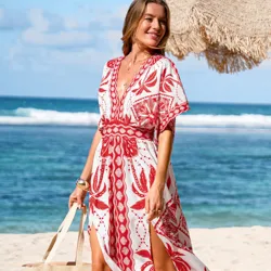 A person on a beach is wearing a white and red patterned dress with short sleeves and a v-neck, holding a light-colored woven tote bag.