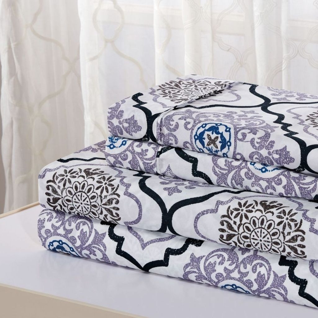 Stack of folded bed sheets featuring a floral and geometric pattern in purple, black, and blue on a white background.