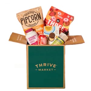 A Thrive Market box containing Pipcorn snacks, SmartyPants vitamins, Three Wishes cereal, Simple Mills crackers, Bare Bell protein bar, Herr's cheese balls, and Mrs. Meyer's hand soap.