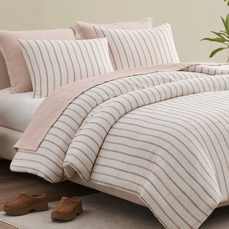 A striped bedding set with matching pillows in soft pink and white tones is displayed on a bed, with brown clogs placed on a light rug nearby.