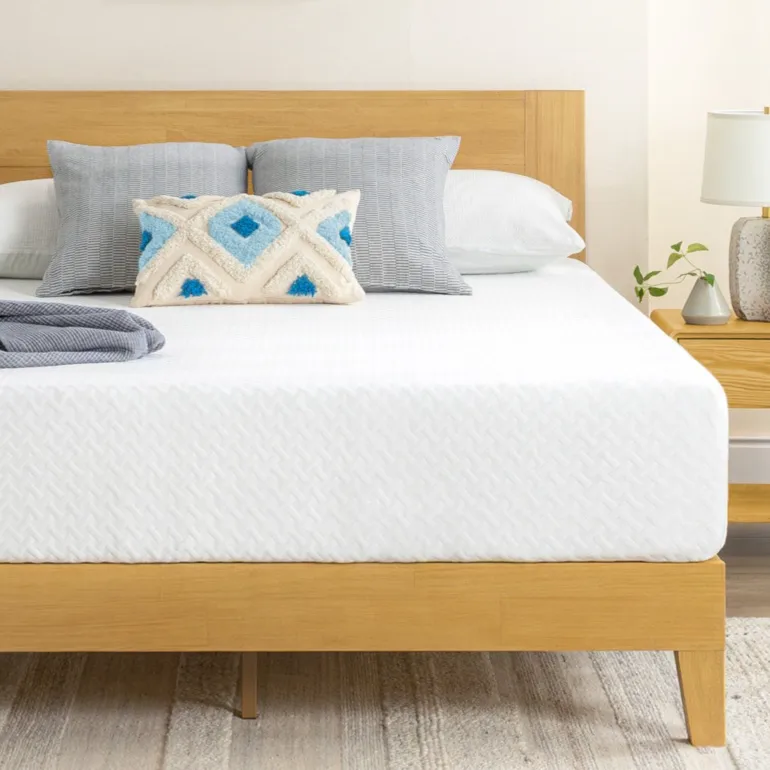 A minimalist bedroom setup featuring a wooden bed frame with a white mattress, gray and patterned pillows, a potted plant, a side table with a lamp, and a neutral rug on a wooden floor.