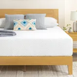 A minimalist bedroom setup featuring a wooden bed frame with a white mattress, gray and patterned pillows, a potted plant, a side table with a lamp, and a neutral rug on a wooden floor.