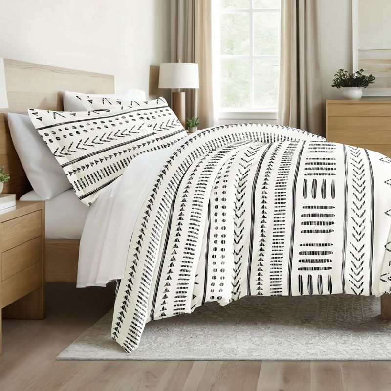 A bed is adorned with a duvet and pillowcases featuring a black and white geometric pattern. The room includes a wooden headboard, side tables, and a window with natural lighting.