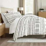 A bed is adorned with a duvet and pillowcases featuring a black and white geometric pattern. The room includes a wooden headboard, side tables, and a window with natural lighting.
