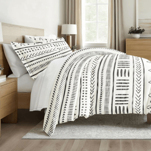 A bed is adorned with a duvet and pillowcases featuring a black and white geometric pattern. The room includes a wooden headboard, side tables, and a window with natural lighting.