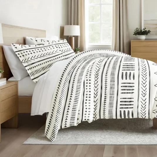 A bed is adorned with a duvet and pillowcases featuring a black and white geometric pattern. The room includes a wooden headboard, side tables, and a window with natural lighting.