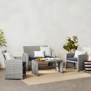 A patio furniture set with a sofa, two chairs, and a coffee table, featuring gray wicker and navy cushions, accompanied by decorative pillows and a small rug.