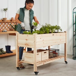 A raised wooden garden bed on wheels with a storage shelf, being used by a person wearing gloves for gardening.