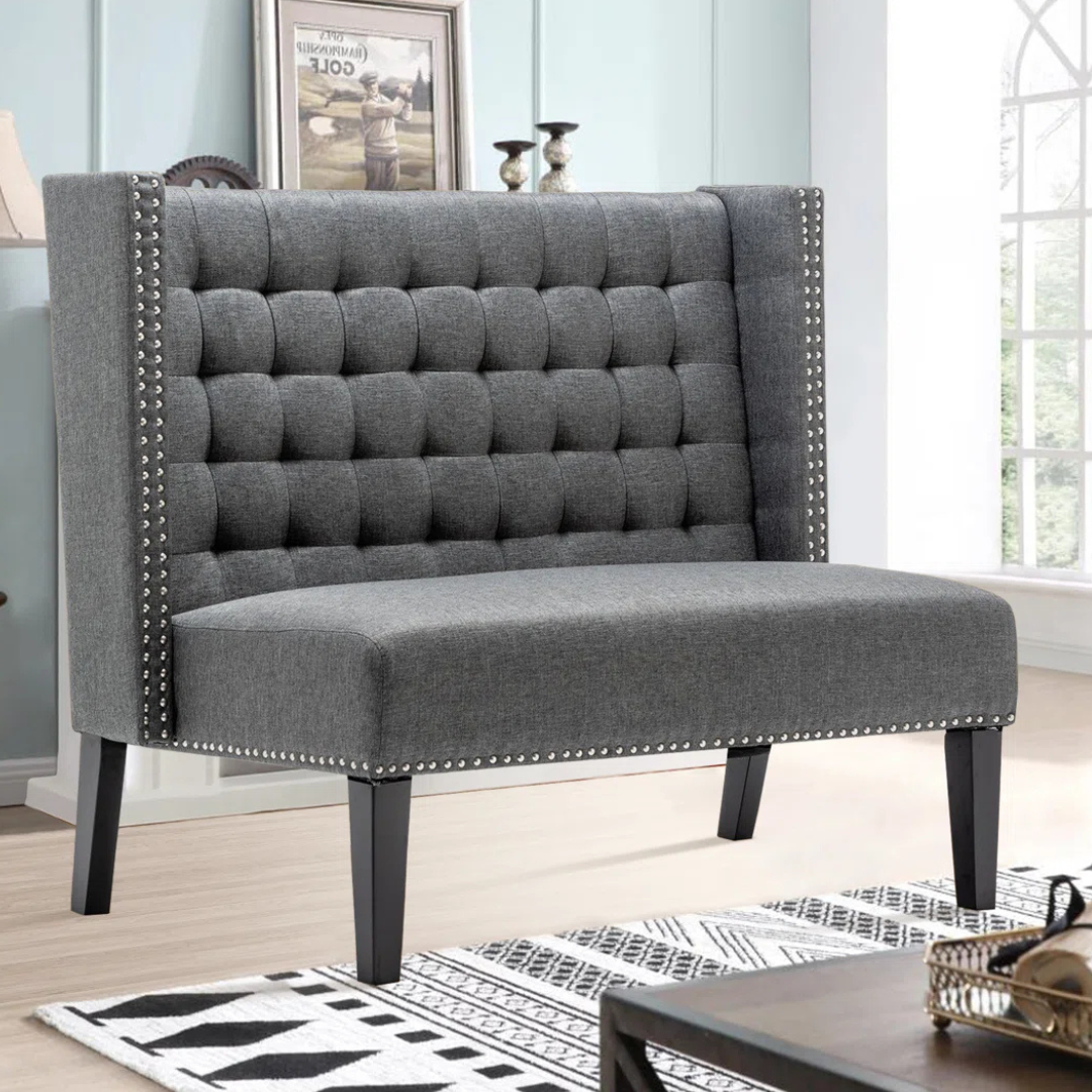 A gray tufted loveseat with studded trim and high backrest, featuring dark wooden legs, in a living room setting with light walls and window.