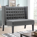 A gray tufted loveseat with studded trim and high backrest, featuring dark wooden legs, in a living room setting with light walls and window.