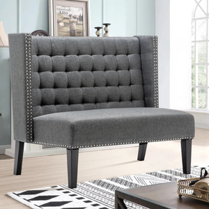 A gray tufted loveseat with studded trim and high backrest, featuring dark wooden legs, in a living room setting with light walls and window.