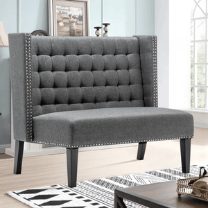 A gray tufted loveseat with studded trim and high backrest, featuring dark wooden legs, in a living room setting with light walls and window.