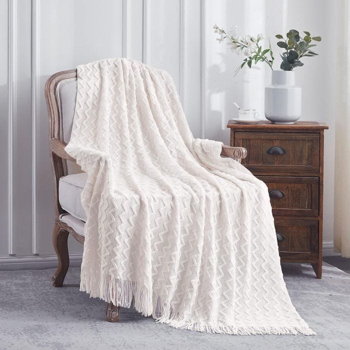 A white, fringed throw blanket with a zigzag pattern is draped over an upholstered chair next to a dark wooden side table. The table holds a vase with flowers and a cup.