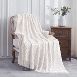 A white, fringed throw blanket with a zigzag pattern is draped over an upholstered chair next to a dark wooden side table. The table holds a vase with flowers and a cup.