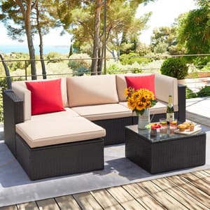A 5-piece sectional conversation set features a dark wicker frame with beige cushions and two red throw pillows. It includes a matching glass-top coffee table adorned with drinks, pastries, and a vase of sunflowers, set on a wooden deck with a scenic background.