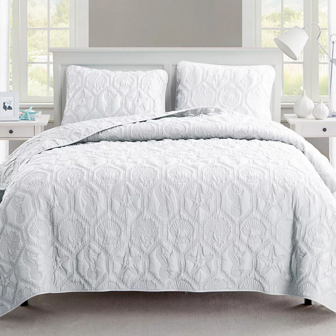 A white quilt with embossed sea-themed patterns including starfish, seashells, and seahorses is displayed on a bed with matching pillow shams.