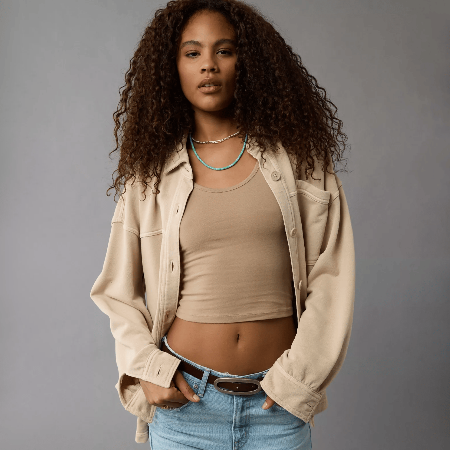 A woman wearing a beige cropped tank top, a loose beige jacket, and blue jeans with a turquoise beaded necklace.