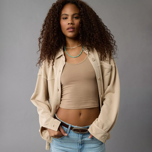 A woman wearing a beige cropped tank top, a loose beige jacket, and blue jeans with a turquoise beaded necklace.