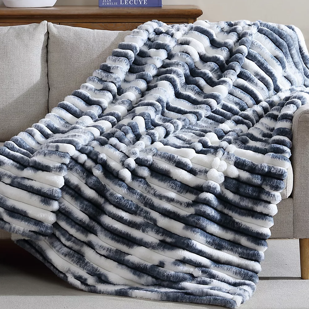 A plush throw with a wavy striped pattern in shades of gray and white, draped over a light-colored sofa, offering a cozy and stylish accent.
