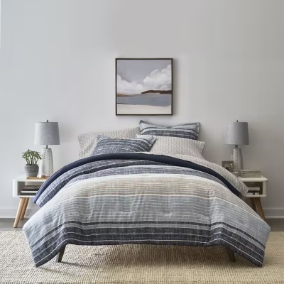 A bedding set with a striped pattern in shades of blue and grey, consisting of a comforter, pillows, and matching sheets, is displayed on a bed with two bedside tables and lamps.