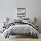A bedding set with a striped pattern in shades of blue and grey, consisting of a comforter, pillows, and matching sheets, is displayed on a bed with two bedside tables and lamps.