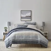 A bedding set with a striped pattern in shades of blue and grey, consisting of a comforter, pillows, and matching sheets, is displayed on a bed with two bedside tables and lamps.