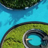 Aerial view of a circular pool area featuring lush green landscaping and blue water, with a person relaxing in a circular section.
