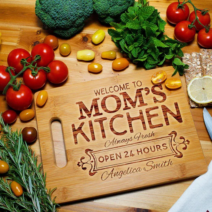 A wooden cutting board engraved with \“Welcome to Mom's Kitchen\“ is surrounded by fresh vegetables like tomatoes, broccoli, and herbs, along with sliced lemon and crackers.