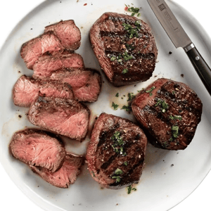 Slices of medium-rare steak with grill marks are presented on a white plate, garnished with chopped herbs. A steak knife is placed beside the steaks.