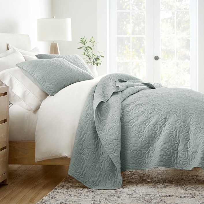 A neatly arranged bed features a light blue quilted comforter with a swirling pattern, matching pillow shams, and a white sheet set. A wooden nightstand with a lamp and a small plant complete the cozy bedroom setting.