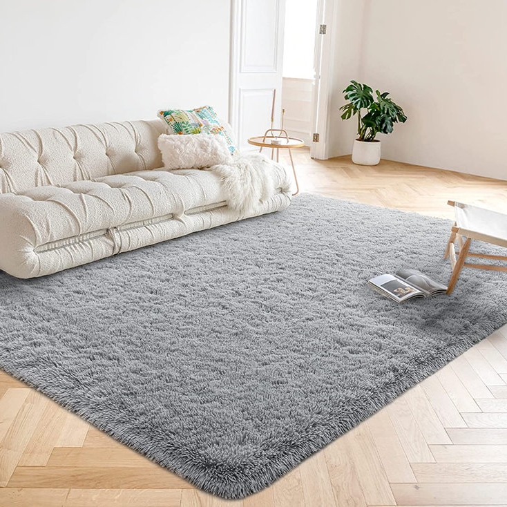 A plush gray area rug is laid out in a living room with a cream sofa, a small side table, and a houseplant in the background.