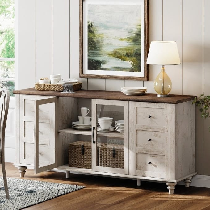 A wooden sideboard with a dark top features glass and solid doors, drawers, and is adorned with a table lamp and artwork. It contains dishware and wicker baskets, set on a wooden floor with a rug nearby.