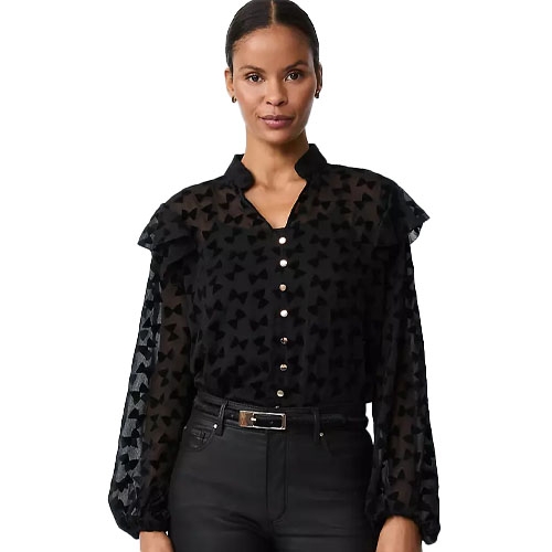 A woman is wearing a sheer black blouse with a pattern of bows, featuring ruffled shoulders, a button-down front, and balloon sleeves, paired with black pants and a belt.