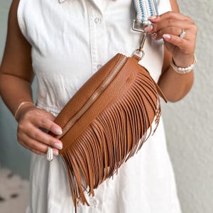 Tan faux-leather crossbody bag with a textured surface, featuring long fringe detail and a single zipper closure. The adjustable strap is striped in blue and white, and the style suits casual or bohemian looks.