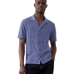 A man is wearing a blue, short-sleeve button-up shirt with a subtle vertical stripe pattern, paired with dark jeans.