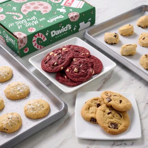 Various types of cookies, including red velvet and chocolate chip, are arranged on plates and baking sheets, with a branded green box featuring holiday-themed designs in the background.