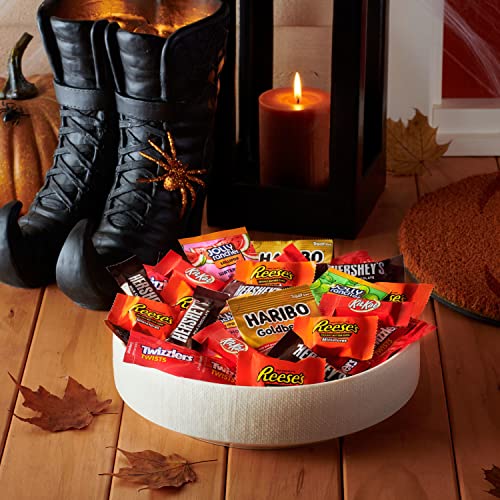 A variety of Hershey's Halloween candy, including Reese's, Twizzlers, and Haribo Goldbears, is displayed on a flat surface, accented by autumn leaves and a candle.