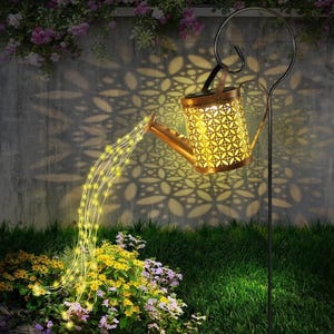 A decorative watering can light in a garden projects intricate patterns on a wall while its spout emits string lights over flowers, creating the illusion of pouring light.