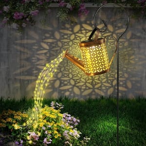 A decorative watering can light in a garden projects intricate patterns on a wall while its spout emits string lights over flowers, creating the illusion of pouring light.
