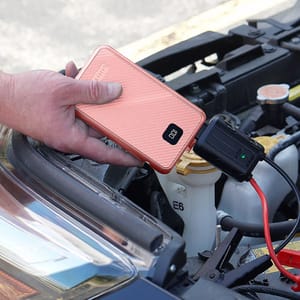 A person is using a pink portable jump starter to charge a car battery, with the device connected via red and black clamps.
