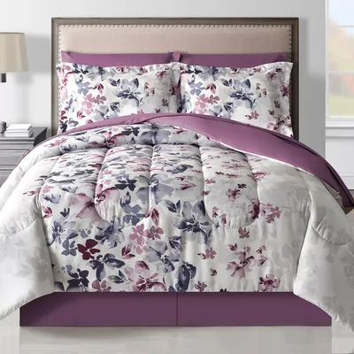 A bed is adorned with a floral-patterned comforter and matching pillow shams in shades of purple and white, accompanied by a upholstered headboard and a side table with a lamp.