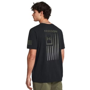 A man wears a black UA Freedom Flag T-Shirt, featuring \“FREEDOM\“ text and a stylized American flag with the Under Armour logo on the back, and an American flag design on the sleeve. The shirt blends athletic style with a patriotic theme.