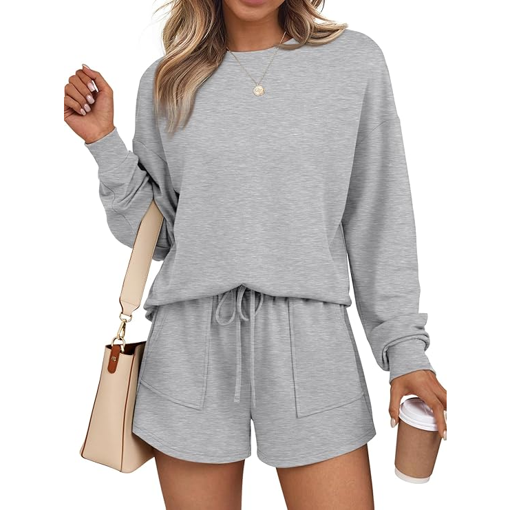 A woman is wearing a gray long-sleeve sweatshirt and matching drawstring shorts. She holds a beige handbag and a coffee cup, accessorized with a delicate necklace.