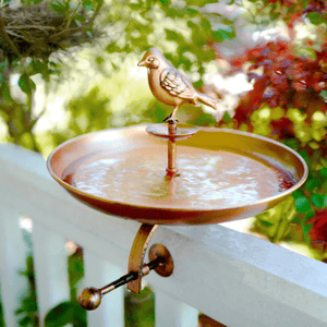 A decorative metal birdbath with an attached bird figurine is mounted on a white fence. The surrounding greenery adds to its outdoor appeal.