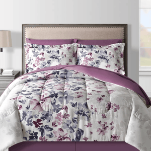 A bed is adorned with a floral-patterned comforter and matching pillow shams in shades of purple, white, and lavender, set against a beige upholstered headboard.