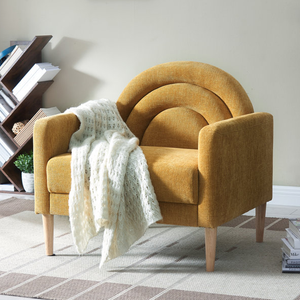 A mustard-yellow contemporary accent chair with a curved backrest, upholstered in soft fabric, and supported by four wooden legs. A white knitted throw drapes over one armrest, set on a checkered rug in front of a modern bookshelf.