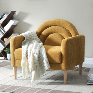 A mustard-yellow contemporary accent chair with a curved backrest, upholstered in soft fabric, and supported by four wooden legs. A white knitted throw drapes over one armrest, set on a checkered rug in front of a modern bookshelf.