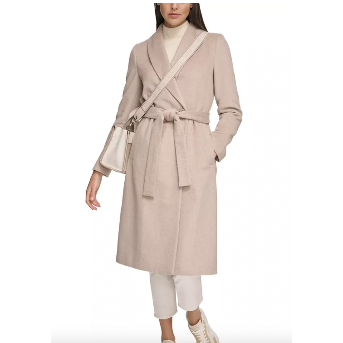 A woman wearing a beige belted coat, white trousers, and white sneakers, carrying a small beige shoulder bag.