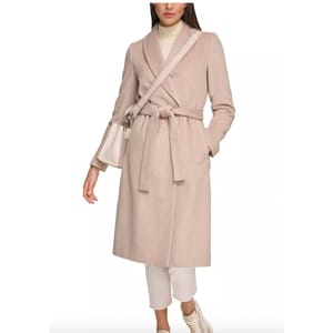 A woman wearing a beige belted coat, white trousers, and white sneakers, carrying a small beige shoulder bag.